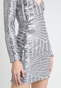 Robe mini en sequins argentés avec manches longues, présentant des motifs rayés horizontaux et diagonaux, une coupe structurée et un tissu texturé.