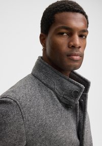 Cappotto grigio a spina di pesce con colletto alto, dotato di chiusura con bottoni e tessuto texturizzato. Il design include dettagli sottili sul colletto.