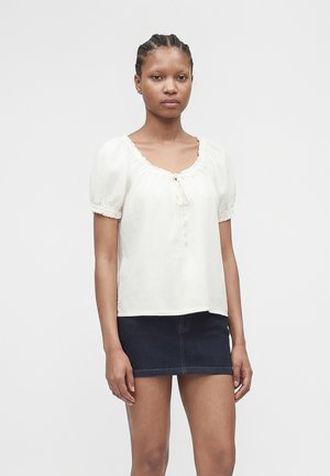 Frau mit geflochtenem Haar, die eine weiße Bluse mit Puffärmeln und Knöpfen sowie einen dunklen Jeans-Minirock trägt, steht vor einem schlichten Hintergrund.