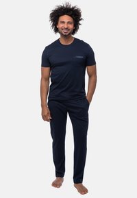 Mörkblå t-shirt med kort ärm och ficka, samt matchande byxor, båda i mjukt tyg. Barfota modell står med händerna i fickorna.