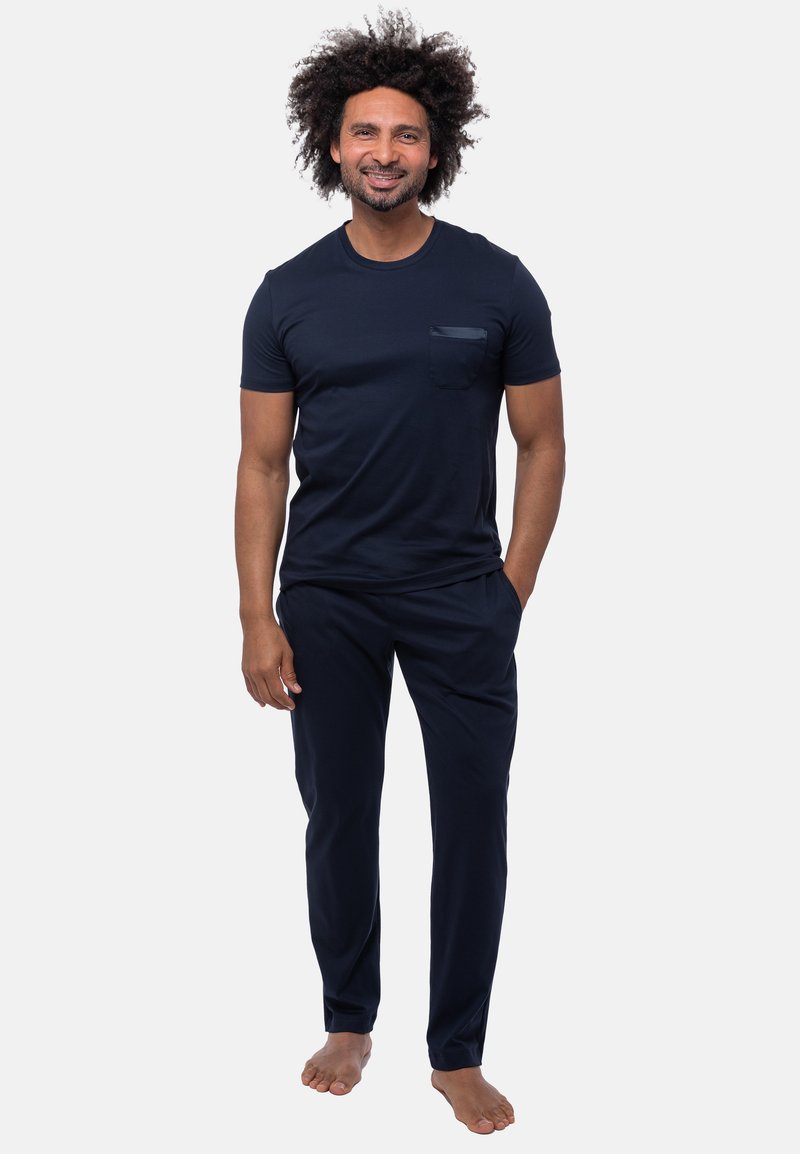 Mörkblå t-shirt med kort ärm och ficka, samt matchande byxor, båda i mjukt tyg. Barfota modell står med händerna i fickorna.