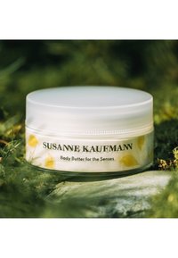 Pot en plastique blanc avec un couvercle lisse, orné d'un motif d'oiseaux jaunes et d'une étiquette en texte noir indiquant "Susanne Kaufmann Beurre corporel pour les sens."