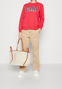 Czerwona bluza z kapturem z logo "GAP", beżowe spodnie w luźnym kroju, białe sneakersy oraz duża torba płócienna z brązowymi skórzanymi uchwytami.