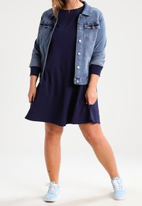 Jeansowa kurtka z dwoma kieszeniami na piersi, krótkie rękawy, jasnoniebieska, na ciemnogranatowej sukience. Model ma na sobie jasnoniebieskie trampki.