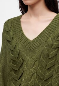 Femme portant un pull en maille vert olive avec col en V et grand motif torsadé texturé.