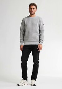 Felpa girocollo grigia in tessuto strutturato, abbinata a pantaloni neri e sneakers bianche, in piedi davanti a uno sfondo neutro.