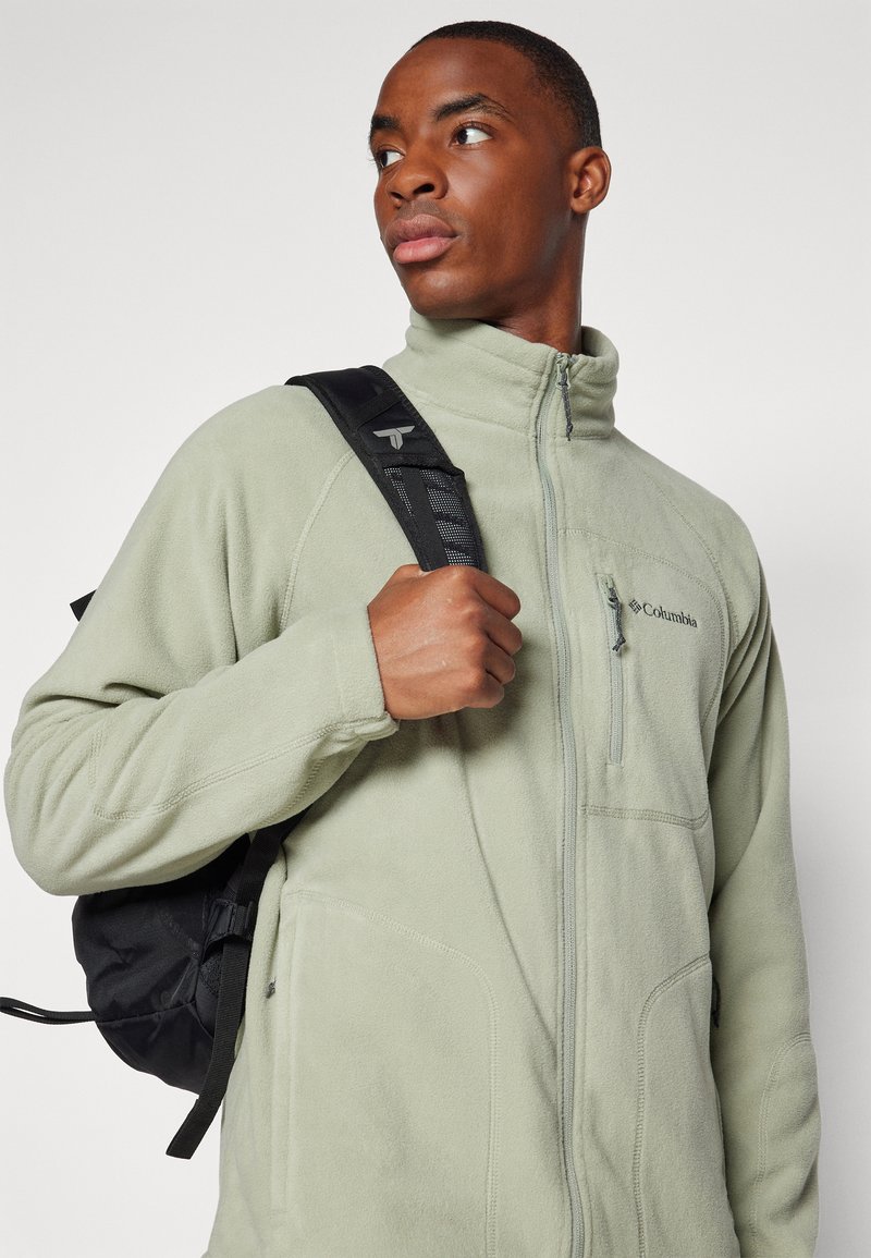 Chaqueta de forro polar verde claro con un collar alto, cremallera frontal y bolsillo en el pecho; presenta detalles bordados y se lleva con una mochila negra.