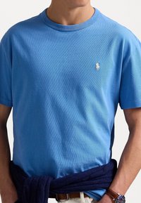 Camiseta de algodón color azul claro con un cuello redondo acanalado y un pequeño logo blanco en el lado izquierdo del pecho, combinada con un suéter azul oscuro atado a la cintura.