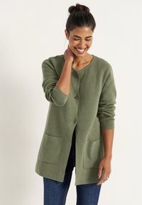Olivgrön cardigan i texturerad tyg, med rund halsringning, knäppning fram och två framfickor. Bärs tillsammans med blå jeans.