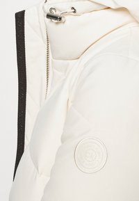 Giacca trapuntata bianca con una zip argentata, cappuccio con coulisse e bordi neri testurizzati. Presenta un logo circolare sulla manica sinistra.