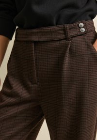 Pantaloni a quadri marroni con vita alta, dotati di due bottoni in vita e tasche laterali. Il materiale ha una consistenza morbida.