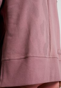 Detailní záběr růžové mikiny s texturovaným maskáčovým vzorem a žebrovaným lemem, zobrazuje část boku nositele.