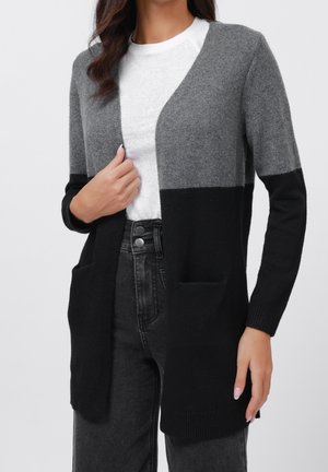 Femme portant un cardigan à manches longues bicolore gris et noir sur une chemise blanche avec un jean noir taille haute, main tenant le revers du cardigan.