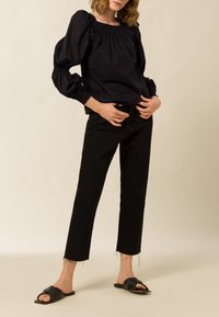 Frau in schwarzer Bluse mit Puffärmeln, hoch taillierten schwarzen Jeans mit fransigen Säumen und schwarzen Riemensandalen, die vor einem schlichten Hintergrund steht.
