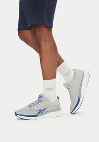 Pantofi sport gri cu accente albastre, material ușor, superior texturat și talpă albă; purtați cu șosete albe care prezintă logo-ul Reebok.