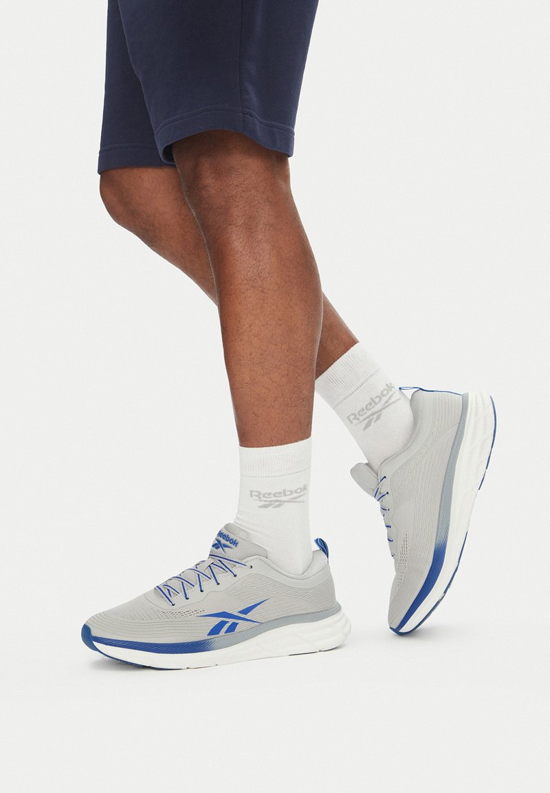 Pantofi sport gri cu accente albastre, material ușor, superior texturat și talpă albă; purtați cu șosete albe care prezintă logo-ul Reebok.