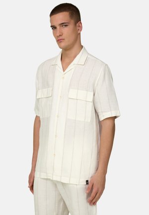 Jeune homme portant une chemise à manches courtes, couleur crème, avec des rayures verticales discrètes et deux poches poitrine, debout devant un fond clair.