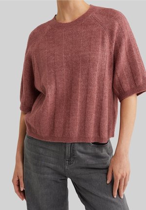 Person trägt einen flauschigen kurzärmeligen Pullover in Mauve mit vertikalen Rippen, kombiniert mit dunkelgrauen Jeans mit hoher Taille vor grauem Hintergrund.
