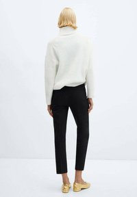 Un pull en maille blanche à col roulé avec des épaules tombantes, associé à un pantalon noir ajusté et des mocassins dorés avec des accents de nœuds. Vue de dos présentée.