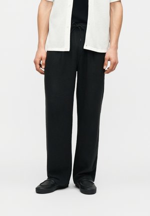 Hombre con pantalones negros sueltos con cintura de cordón, zapatos negros y una camisa blanca de manga corta abierta sobre una camiseta negra, de pie sobre un suelo blanco.