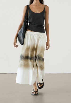 Femme portant un haut noir sans manches, un pantalon ample beige et noir tie-dye, des sandales noires, et tenant un grand sac à bandoulière noir tissé.