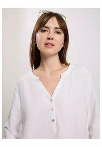 Biała bluzka z guzikami i dekoltem w kształcie litery V, z trzema guzikami i długimi rękawami. Tkanina wydaje się lekka i gładka.