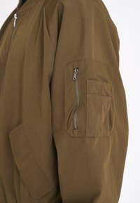 Brauner Jackenärmel mit Reißverschlusstasche und zwei kleinen eingenähten Fächern, dargestellt auf weißem Hintergrund.