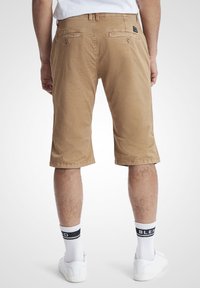 Beige shorts laget av bomull, kne-lange, med løs passform. Har to baklommer og en sydd logo-lapp nær midjebåndet.