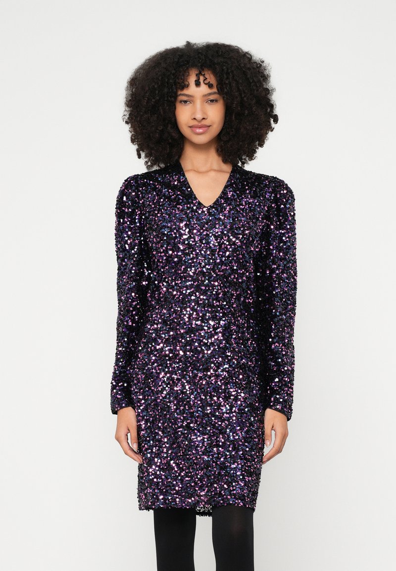 Γυναίκα με σγουρά μαλλιά, φορεμένη με μακρυμάνικο, μέχρι το γόνατο φόρεμα καλυμμένο με μωβ και μαύρα παγιέτες, στέκεται μπροστά σε απλό φόντο.