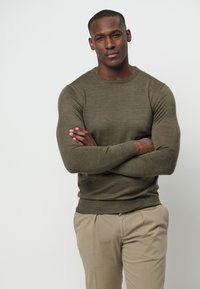 Suéter de punto verde oliva con mangas largas, cuello redondo y puños acanalados, combinado con pantalones beiges. Textura suave, diseño ajustado.