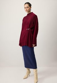 Bordo ohlapen pulover iz pletenine z visokim ovratnikom, okrašen s srebrno zaponko za varovanje. Kombiniran z mornarsko modro midi krilo in kremnimi škornji z visoko peto.