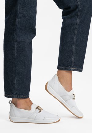 White slip-on shoes with a textured fabric upper, a buckle accent, and a rubber sole. Paired with dark blue denim pants.