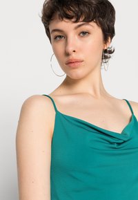 Camisola de cetim teal com alças finas, apresentando um decote drapeado e uma textura suave. Modelo a usar grandes brincos de argola.