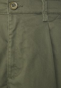 Olivově zelené kalhoty z bavlny, s velkou přední kapsou, zapínáním na knoflíky a viditelnými švy. Hladký povrch s jemným leskem.