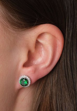 Smaragd-Ohrstecker, die in einer kreisförmigen Silberfassung sitzen, umgeben von kleinen klaren Kristallen. Die Ohrringe liegen an einem menschlichen Ohr.