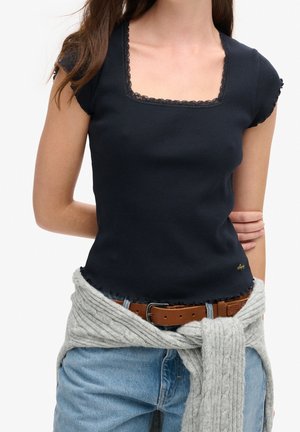 Kobieta w czarnej prążkowanej bluzce z kwadratowym dekoltem i koronkową lamówką, jasnoniebieskich jeansach z brązowym paskiem oraz szarym dzianinowym swetrze związanym w pasie.