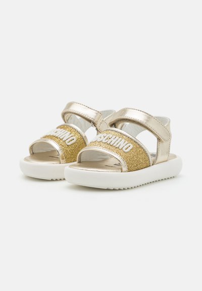 Zelta metāla sandales ar mirdzošu zelta siksnu, kurā ir baltā krāsā rakstīts "MOSCHINO", baltām zoles un regulējamām velcro sprādzēm.