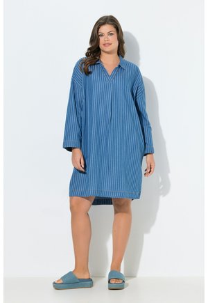 Donna con un abito blu a righe sottili, ampio e a maniche lunghe, e sandali blu senza lacci, in piedi su sfondo bianco.