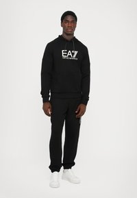 Set di felpa nera con cappuccio e pantaloni jogger realizzati in tessuto morbido. La felpa presenta una tasca frontale e una stampa del logo, abbinata a sneakers bianche.