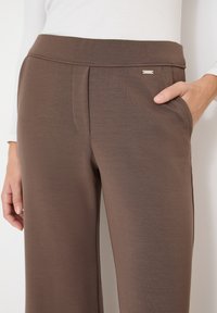 Pantalon marron taille haute avec poches latérales, porté avec un haut blanc à manches longues ; une main reposant dans la poche.