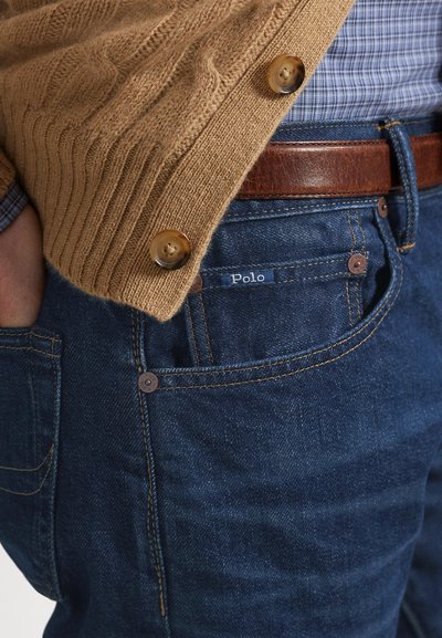 Zbliżenie osoby noszącej niebieskie dżinsy z metką Polo, brązowy skórzany pasek, niebieską koszulę w kratę oraz brązowy dziany kardigan z guzikami.