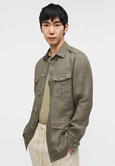 Πουκάμισο-σακάκι από λινό σε ελαιώδες πράσινο χρώμα με κουμπιά, δύο τσέπες στο στήθος και άνετη γραμμή, φορεμένο πάνω από ανοιχτό πράσινο τοπ.