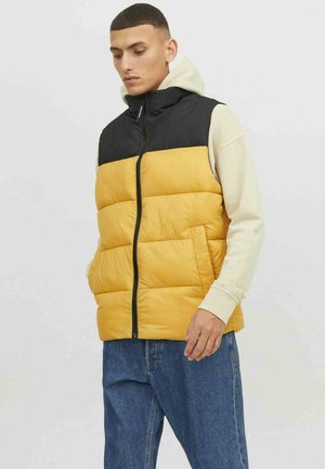 Yellow and black puffer vest with zip closure, featuring a quilted design, high collar, and pockets, worn over a light hoodie and blue jeans.