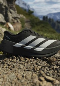 Черна маратонка Adidas Terrex с бели ивици на скалиста планинска пътека под ясен небосвод.