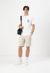 Witte t-shirt met "LS" geborduurd, beige shorts en witte sneakers. Model houdt een zwarte schoudertas met blauwe accenten vast. Eenvoudige achtergrond.