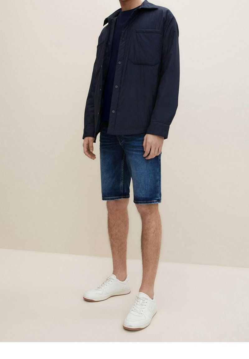 Navy button-up jacket with a pocket, navy t-shirt underneath, denim shorts, and white sneakers with textured soles.