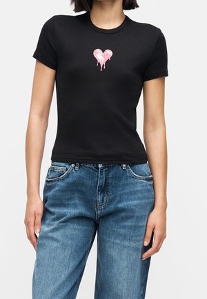 Femme portant un t-shirt noir ajusté avec un petit motif de cœur rose en train de fondre sur la poitrine, et un jean bleu sur un fond uni.
