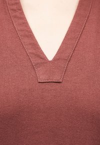 Κοντινό πλάνο φωτεινής επιδερμίδας που φοράει τοπ από υφασμάτινη ύλη σε σκουροκόκκινο χρώμα με υφή, με V-σχήμα στο λαιμό και ορατή ραφή.