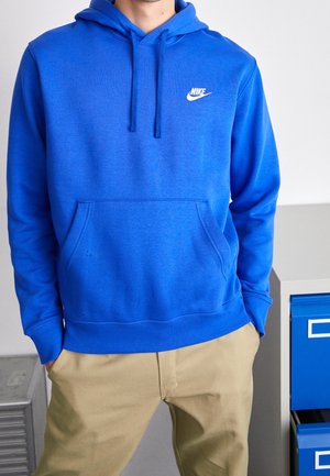Person iført en knaldblå Nike-hættetrøje med beige bukser, hænderne i lommerne, står ved siden af et gråt metalskab med blå skuffer.