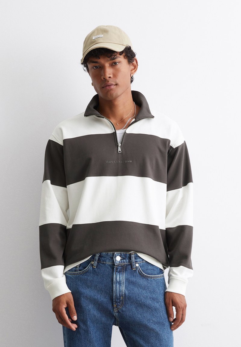 Barna és fehér csíkos pulóver, cipzáras gallérral. A mellkason logóval és karakteres anyaggal rendelkezik. Kék farmerrel párosítva. Bézs sapka a fején.